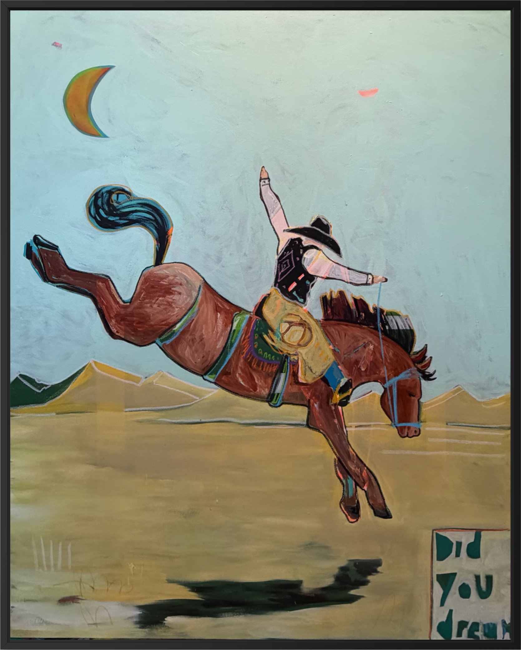 A framed painting depicting a cowboy riding a bucking brown horse across a desert landscape under a pale sky with a crescent moon.